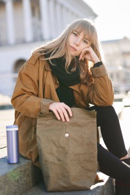 Ceketli genç sarışın kız beslenme çantasıyla dışarıda kameraya bakıyor. Öğle yemeği konsepti.