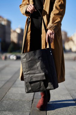 Siyah ekolojik kağıt, mavi gökyüzü ve büyük şehir arka planına sahip turuncu siper giyen bir kızın ellerinde torba yaptı. Moda eko kese kağıdı konsepti.