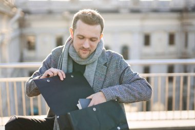 Güneşli bir günde şehir manzaralı siyah sırt çantasından not defteri alan gülümseyen genç adam. Pozitif düşünce konsepti