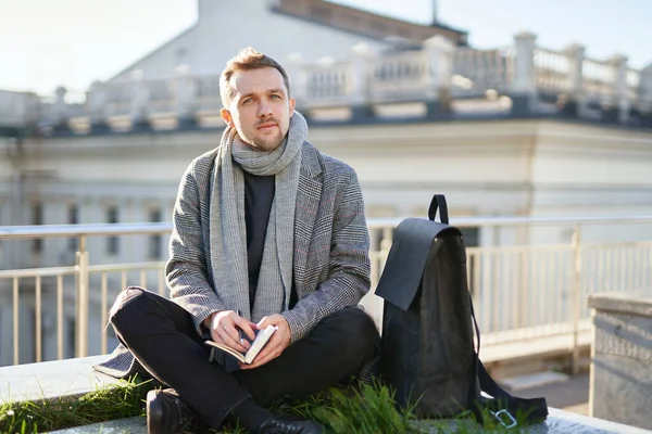 Gri atkı ve ceketli genç adam elinde not defteri ve kalemle çatıda oturuyor ve şehir manzaralı arka planda düşünüyor. Çalışma veya öğrenim kavramı.
