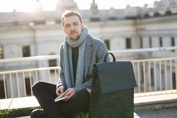 Eşarplı ve ceketli genç çekici bir adam kameraya bakıyor, notla oturuyor. Güneşli bir günde açık hava konseptini çalışmak ya da öğrenmek