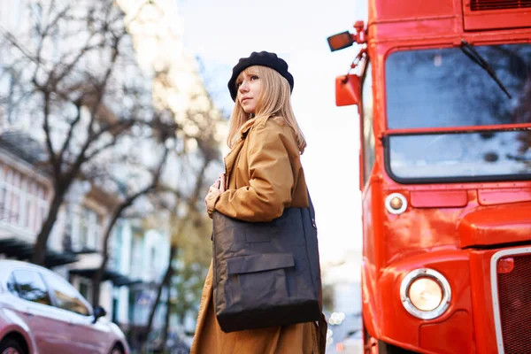 Siyah bereli ve kırmızı otobüslü turuncu siperli çekici bir genç kadın. Çift katlı arka planda siyah çanta tutan mutlu insan. Pozitif düşünce ya da seyahat kavramı