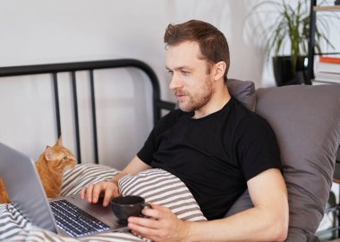 Genç beyaz bir adam laptopta çalışırken sabah kahvesini içiyor, battaniyenin altında yatıp video konuşması yapıyor. Genç adam ya sınava hazırlanıyor ya da internette sörf yapmak için dizüstü bilgisayar kullanıyor