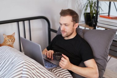 Sakallı genç adam battaniyenin altındaki yatakta dizüstü bilgisayarla çalışıyor ve yanında bir kedi yatıyor. Uzak çalışma veya çevrimiçi çalışma kavramı