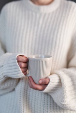 Bir bardak sıcak içecekle kadın eli yakın çekim. Sabahın köründe elinde çay ya da kahve tutan beyaz kazaklı güzel bir kız. Tasarımın için boş bardak. Kış konforu, sabah vakti