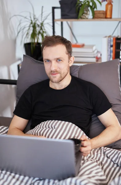 Young bearded man is working on a laptop on the bed covered with blanket. Remote work, online studying or surfing the web concept