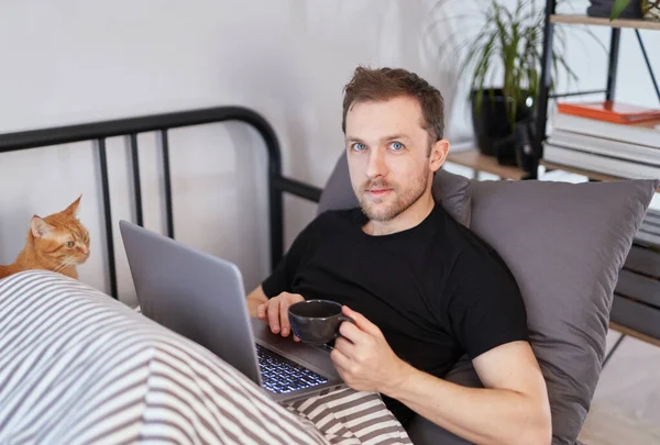 티셔츠를 매력적 베어링을 남자가 자신의 사무실 침대에 노트북을 고양이와 카메라를 — 스톡 사진