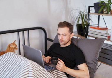 Sakallı iş adamı yanında kedi yatarken battaniyenin altında dizüstü bilgisayarla çalışıyor. Uzak çalışma, çevrimiçi öğrenim veya alışveriş kavramı