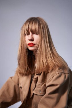 Parlak makyajlı genç sarışın kadın portresi. Dudaklarında parlak kırmızı ruj olan güzel bir sarışın. Uzun sarı saçlı, bej ceketli güzel bir kız.