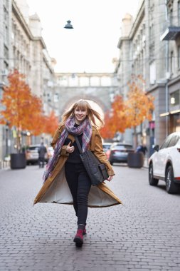 Uzun turuncu trençkot giyen, siyah kotlu ve atkılı, Avrupa şehrinin caddesinde yürüyen çekici sarışın kadının moda tarzı fotoğrafı.