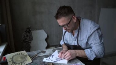 Linocut sanatçısı el yapımı baskı işi. Önlüklü, sakallı orta yaşlı bir adam kesici alet kullanarak linocut yapıyor. 4k görüntü