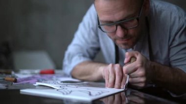 Linocut sanatçısı elle oyma işlemi yapıyor. El yapımı iş. Önlüklü, sakallı orta yaşlı bir adam kesici alet kullanarak linocut yapıyor. 4k görüntü