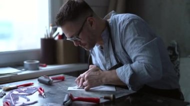 Lithograph sanatçısı el yapımı işçi. Sakallı, orta yaşlı, önlüklü ve gözlüklü bir adam kesme aletini kullanarak linocut yapıyor. 4k görüntü