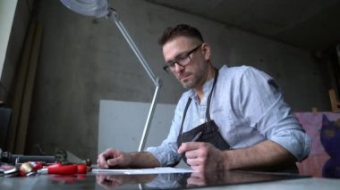 El yapımı pul yapan bir taslak sanatçısı. Linocut sanatçısı baskı imalatı işi. Önlüklü sakallı, orta yaşlı bir adam muşamba üzerinde resim çiziyor. 4k görüntü