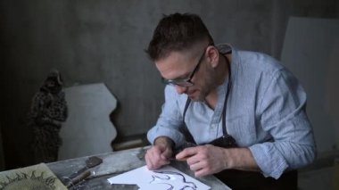 Lithograph sanatçısı el yapımı işçi. Sakallı, önlüklü ve gözlüklü çekici bir erkek kesici alet kullanarak linocut yapıyor. 4k görüntü