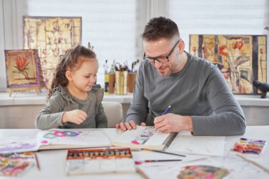 Baba, kızına pastel boyalarla resim çizmesinde yardım ediyor.