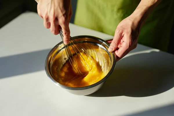 Шеф-повар в зеленом фартуке смешивает яйца в миске с помощью проволочного венчика — стоковое фото
