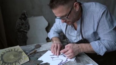 Önlüklü sakallı bir zanaatkar kesme aletleriyle linocut yapıyor.