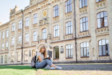 Üniversite kampüsündeki öğrenci yeşil çimlerde dizüstü bilgisayar kullanarak oturuyor.