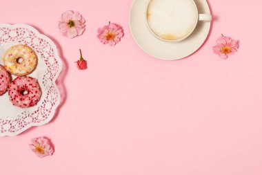 Bir fincan kapuçino, pembe arka planda donut ve güller.