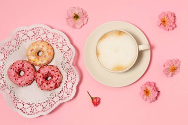 Bir fincan kapuçino, pembe arka planda donut ve güller.