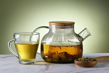 Yeşil zemin üzerinde bitki çayı olan şeffaf çaydanlık ve fincan.