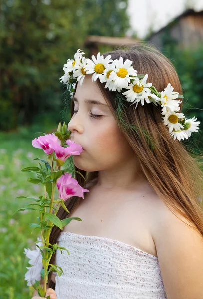 Retrato de uma menina em uma coroa de camomilas em sua cabeça, que está cheirando uma flor — Fotografia de Stock