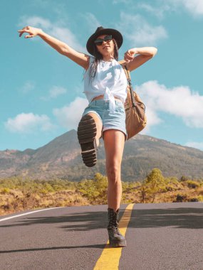 Sırt çantalı kız, bir volkanın ve dağların arka planında yol boyunca yürüyor. Seyahat kavramı