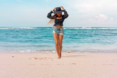 Şapkalı genç bir kadın denizin arka planında sahil boyunca yürüyor. Dinlenme havası, tatil.