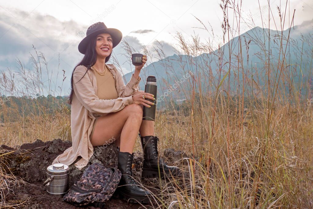 A young woman drinks tea from a thermos against the backdrop of the savannah and mountains. Safari style in clothing. Travel, camping.