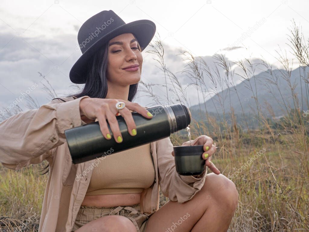 A young woman drinks tea from a thermos against the backdrop of the savannah and mountains. Safari style in clothing. Travel, camping.