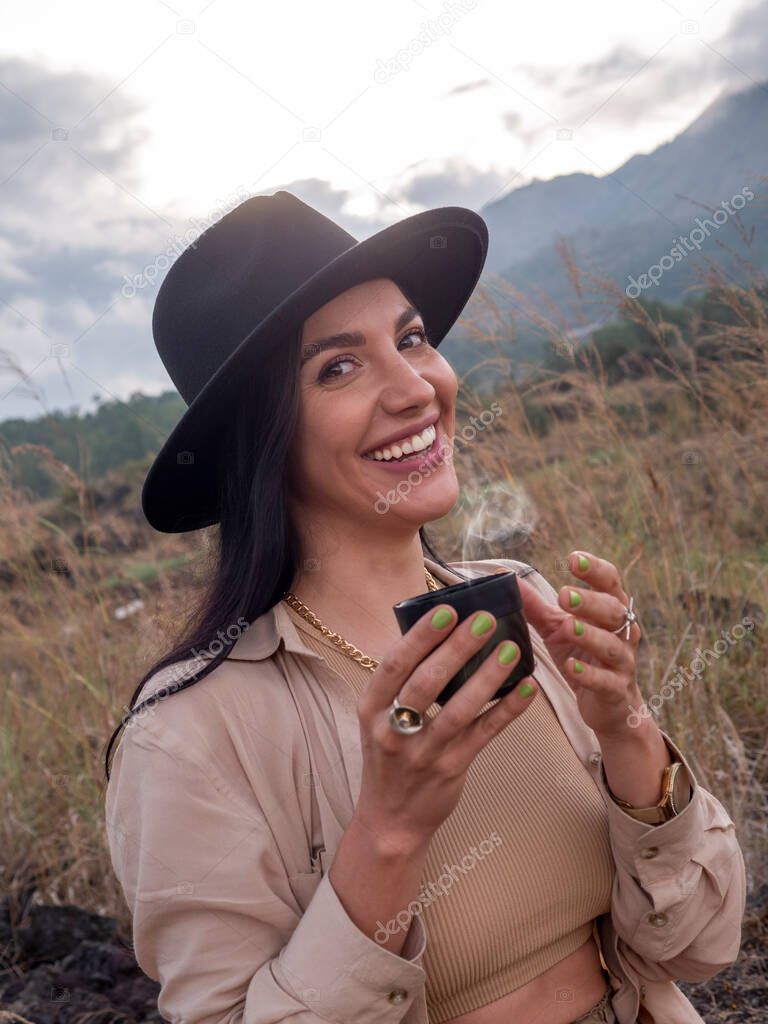 A young woman drinks tea from a thermos against the backdrop of the savannah and mountains. Safari style in clothing. Travel, camping.