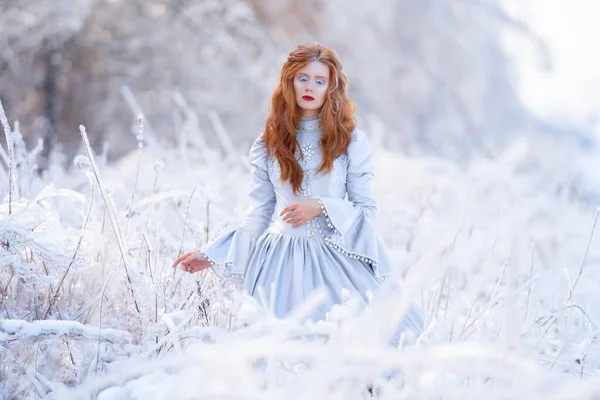 Mulher ruiva jovem, uma princesa, caminha em uma floresta de inverno em um vestido azul. — Fotografia de Stock