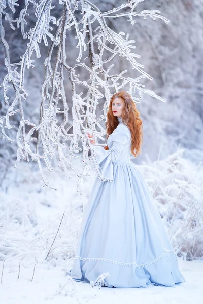 Mulher ruiva jovem, uma princesa, caminha em uma floresta de inverno em um vestido azul. — Fotografia de Stock