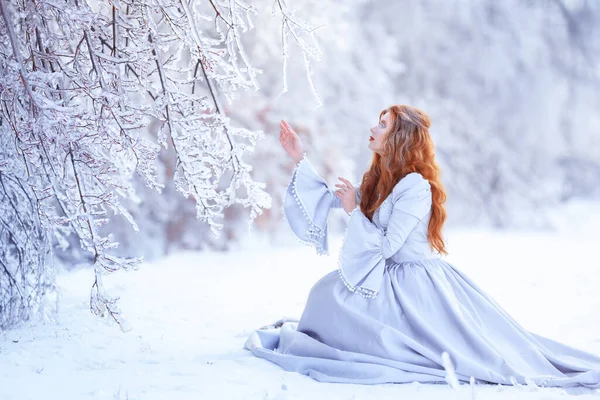 Mulher ruiva jovem, uma princesa, caminha em uma floresta de inverno em um vestido azul. — Fotografia de Stock