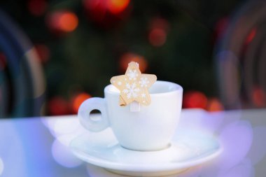 Beyaz porselen kahve fincanının üzerinde Violet Bokeh ve çay tabağı ve kar taneli ahşap dekoratif yıldız. Arka planda bulanık sandalyeler ve kırmızı topları olan Noel ağacı. Yeni yıl kartpostalı. Tebrikler..