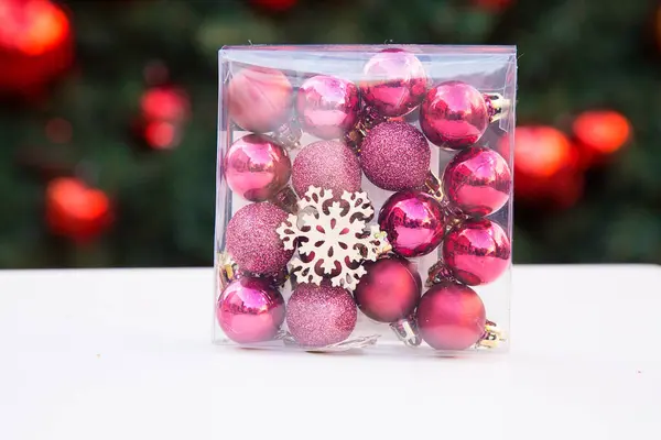 Plastic transparent square box with rose and purple small Christmas balls and wooden snowflake inside. White table. New year tree with red decorations in background. Xmas toys. Preparation for — Stock Photo, Image