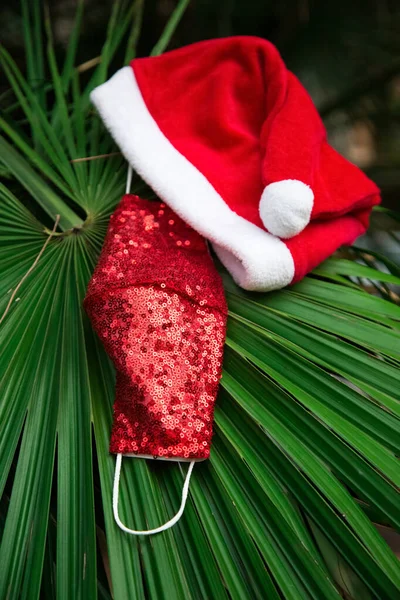 Beyaz kenarlı kırmızı Noel şapkası, yeşil palmiye yaprağında kırmızı koruyucu tıbbi ışıltılı maske. 2021 yılbaşı partisi için aksesuarlar. Tekrar kullanılabilir kovid koruma. Noel arifesi için güvenlik kiti. Yeni gerçeklik