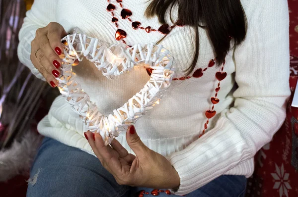 Woman in white sweater holds in hands white wooden heart with electric garland. New year decoration. Fingers with red nails. Christmas clothes. Tender and love concept. Saint Valentains day — Stock Photo, Image