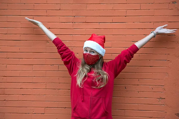 Noel kostümü giymiş sarışın kız ve kırmızı koruyucu maske elleri havada duruyor. Hoş geldiniz ve neşeli bir jest. Beyaz kadın, yeni yıl korumasında. Arkadaki turuncu tuğla duvar, boşluğu kopyala. 