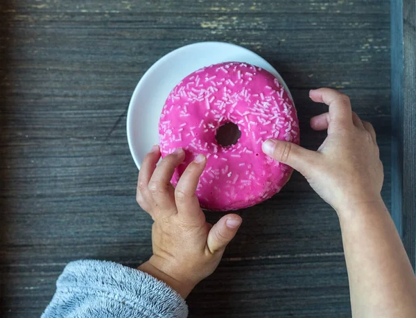 Baby takes bright pink donut from white plate. Childs hands. Grey wooden background. Sweets for kids breakfast. Traditional American meal. Top view to a table with round objects. Gluten products