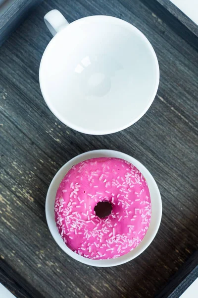 Vertical picture of bright pink donut on white round plate and tea cup above. Aged wooden background. Isolated objects. Traditional American breakfast. Copy space. Geometrical concept. Eating at home