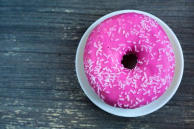 Beyaz yuvarlak tabakta parlak pembe çöreğe ve eskimiş ahşap arka planda eski ahşap çöreğe. İzole bir nesne yaklaşıyor. Kahvaltıda geleneksel Amerikan tatlıları. Uzayı kopyala Üstte beyaz şeker süslemesi. 