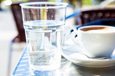 Şeffaf plastik bardak ve bir bardak kapuçino ve arka plandaki kafe masaları bulanıklaştırılmış. Geleneksel İtalyan tarzı kahve servisi. Hala kafeine karşı su içiyor. Sağlıklı diyet. 