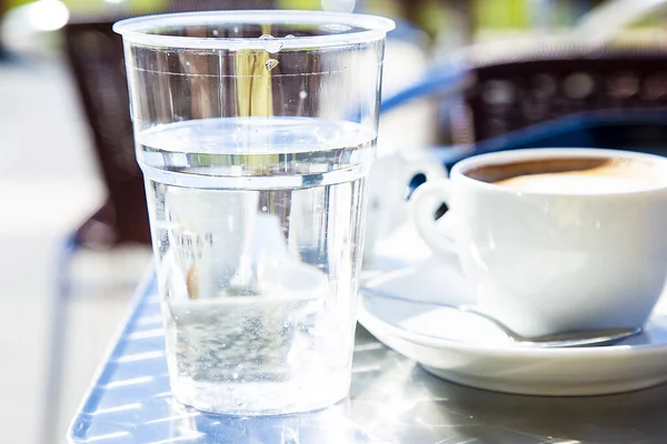 Şeffaf plastik bardak ve bir bardak kapuçino ve arka plandaki kafe masaları bulanıklaştırılmış. Geleneksel İtalyan tarzı kahve servisi. Hala kafeine karşı su içiyor. Sağlıklı diyet. 