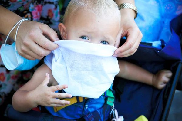 Blond blue eyes baby looks straight. Female hands with surgery mask put white scarf as a mask on childs face. Pandemic contest. No masks for children. Virus protection for infants. Childhood
