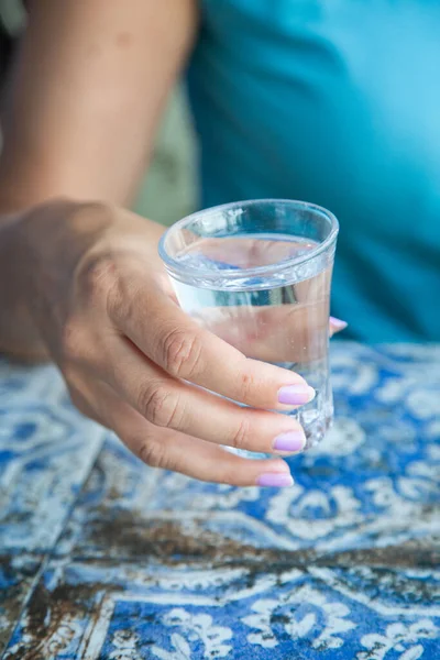 Yakın plan su bardağı, manikürlü çiviler, turkuaz kaplama, desenli fayans masa örtüsü, yumuşak güneş ışığı, rahat bir jest.