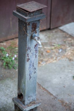 Beton üzerinde paslı metal su musluğu, paslanmış kare sütun, mavi boya, damlayan kapakçık ve tabanda küçük otlar.,