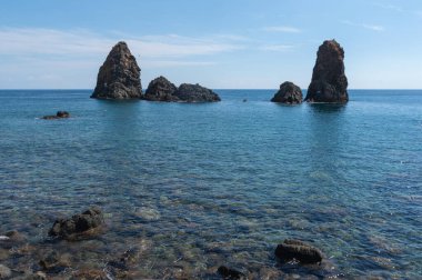 Sicilya 'daki Aci Trezza kıyısındaki berrak mavi su, oradaki bazı volkanik deniz yığınlarına hayranlık duyulur.