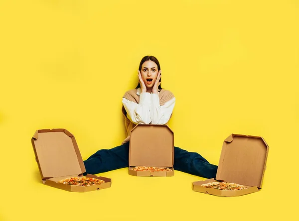 Emotioneel verrast jonge vrouw omringd door drie pizza 's in dozen op gele achtergrond. Pizza-leveringsconcept. Stockfoto